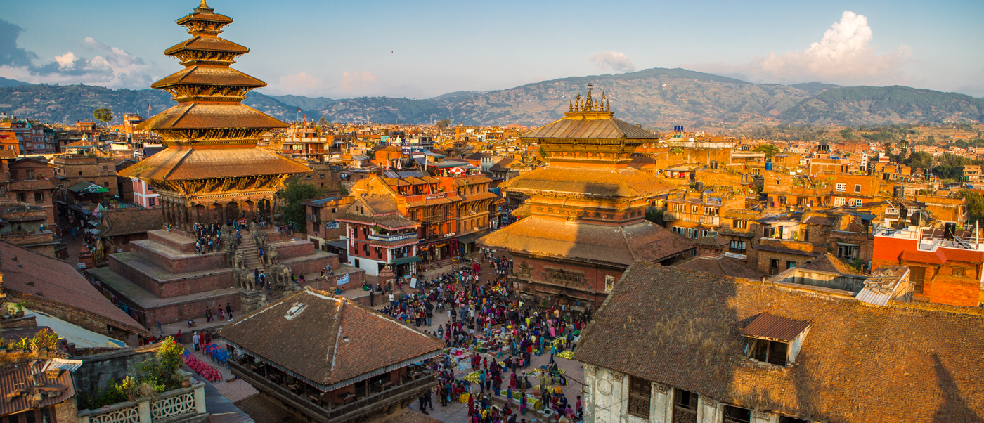 Crowds gather in Kathmandu's historic Durbar Square, surrounded by ornate tiered temples and distant mountains.