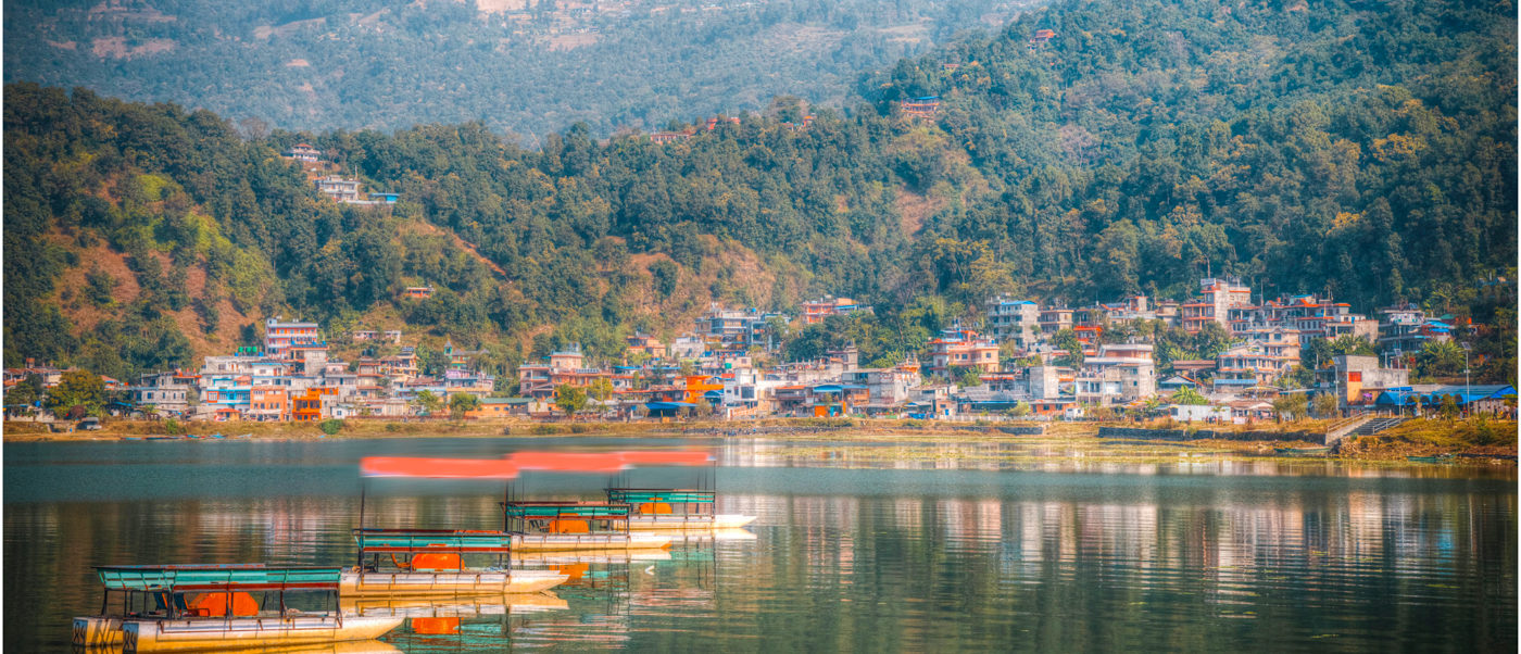Colorful boats float on serene Phewa Lake, reflecting the vibrant town of Pokhara and surrounding green hills.