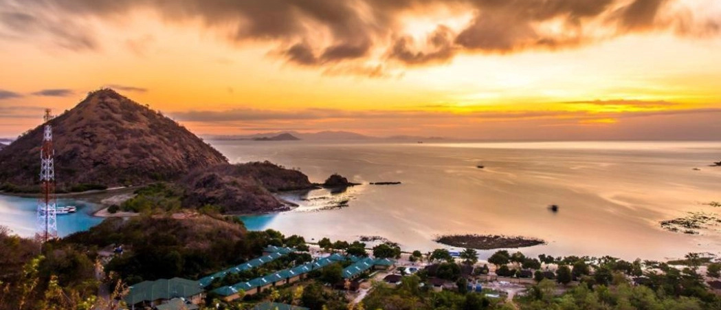 Vibrant golden sunset over Labuan Bajo bay, with a large island and coastal buildings.