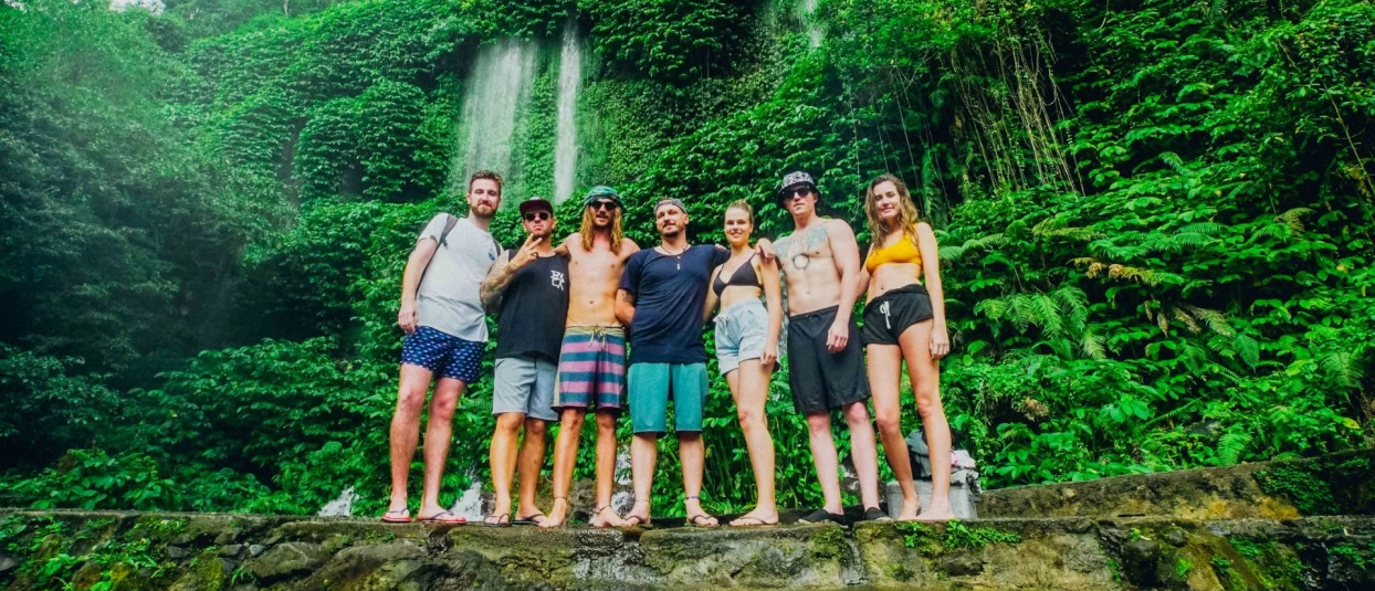 A group of seven young backpackers smiling in front of lush green waterfalls in Lombok.