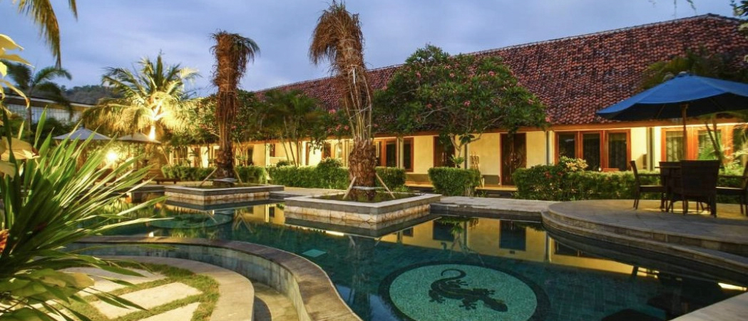 An inviting resort pool with a gecko mosaic, palm trees, and illuminated buildings at dusk in Lombok.