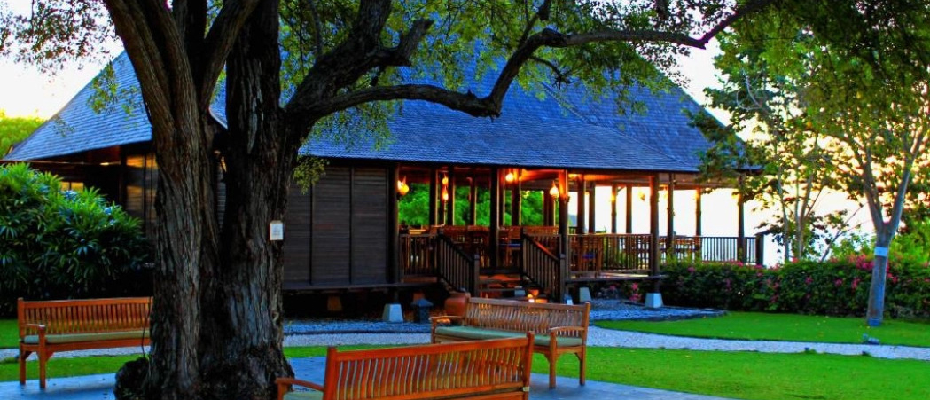 A traditional wooden building with warm lights at dusk, framed by a large tree on Sumbawa island.