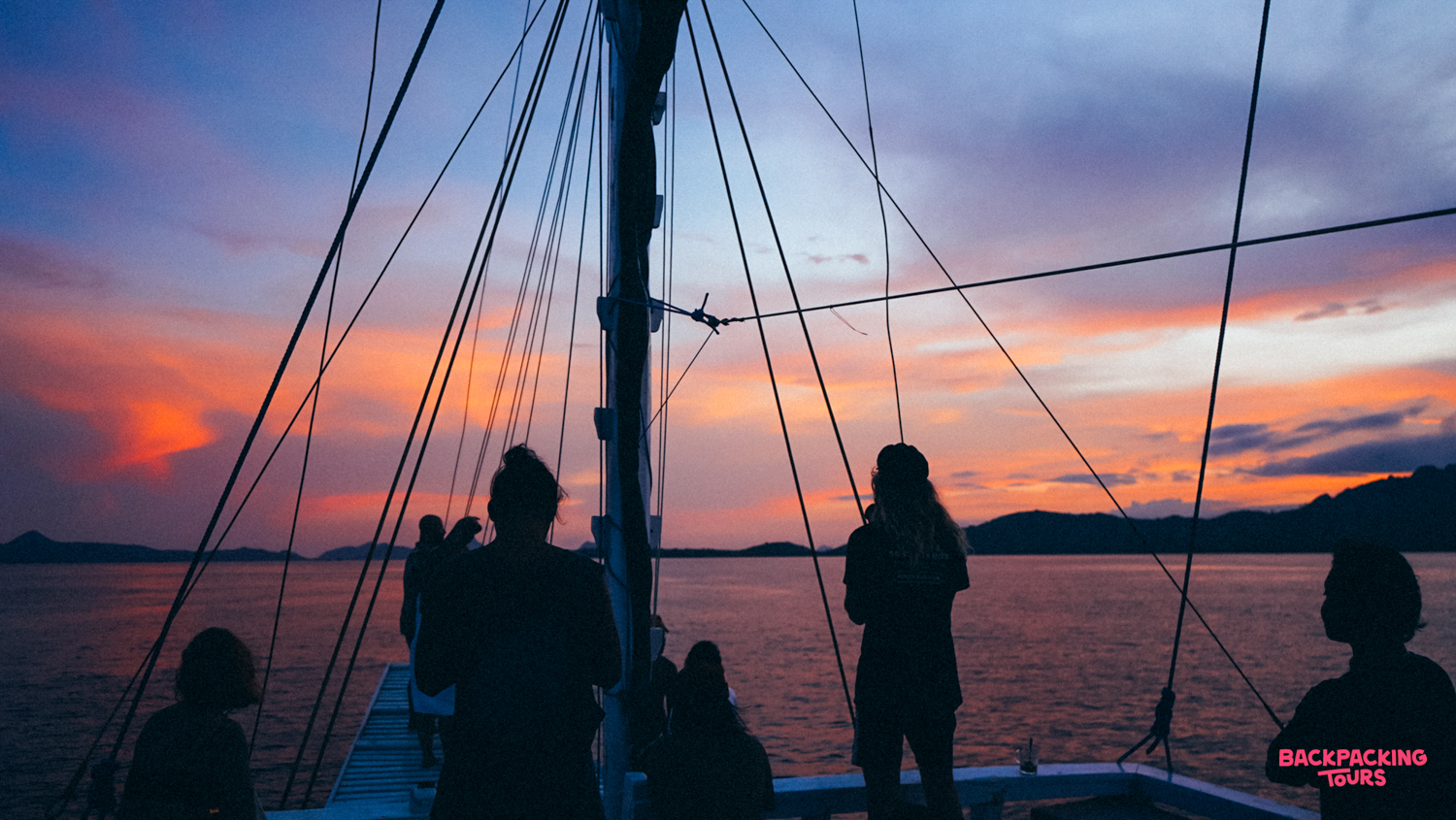 Travelers on a traditional boat watch a vibrant Indonesian sunset over the calm sea.