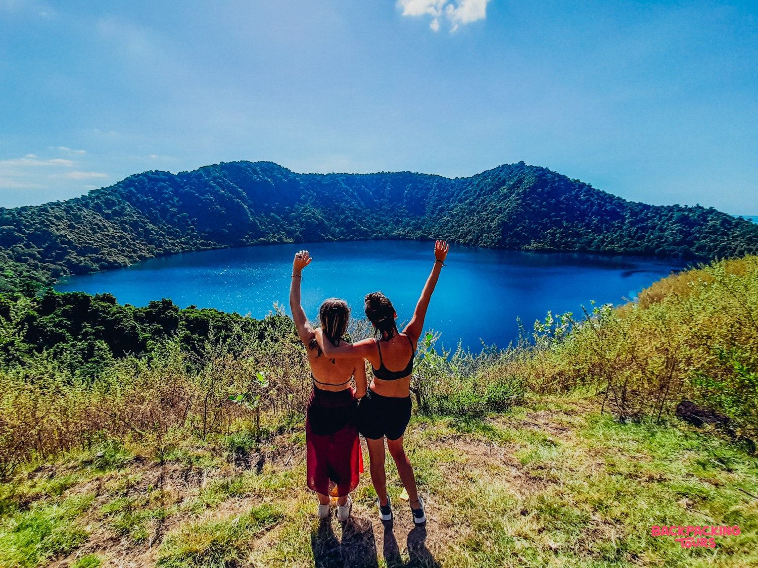 Joyful travelers with raised arms admiring the breathtaking blue crater lake on Satonda Island.