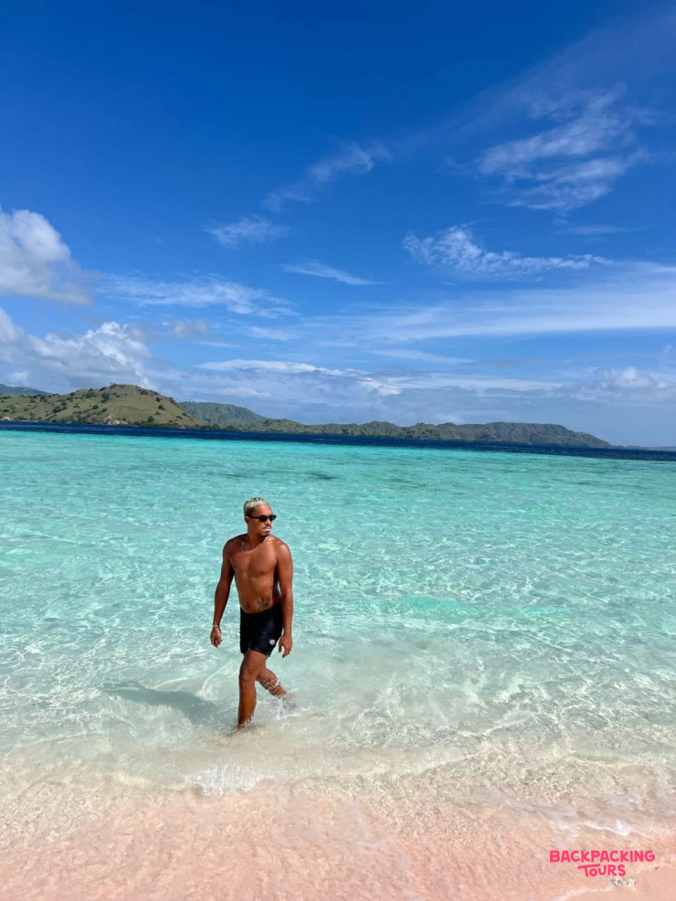 A man strolls through the shallow, clear turquoise water of a beautiful pink sand beach.