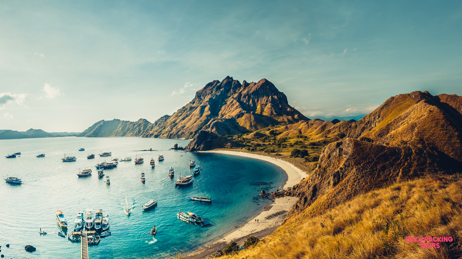 Boats dot a beautiful bay with a sandy beach and rugged hills in Komodo National Park.