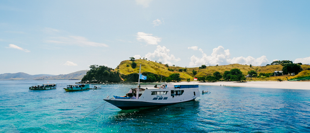 A white liveaboard boat floats on clear turquoise water near Satonda Island's white sand beach.