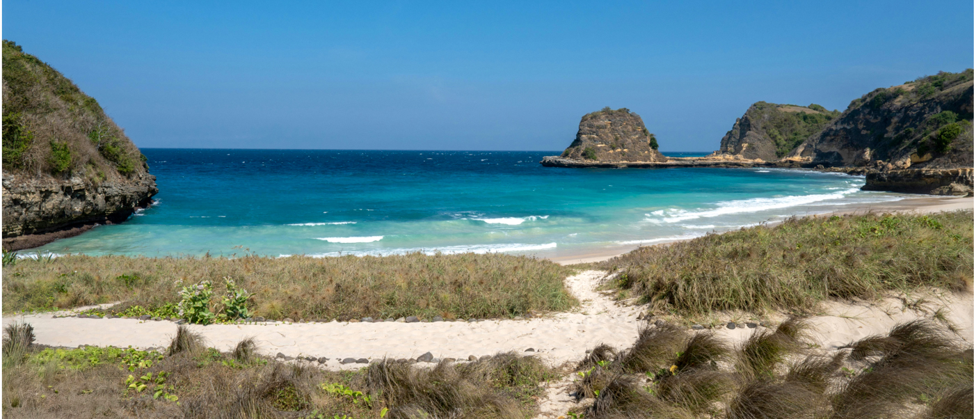 A pristine turquoise bay with a sandy beach and rocky cliffs under a clear blue sky in Lombok.