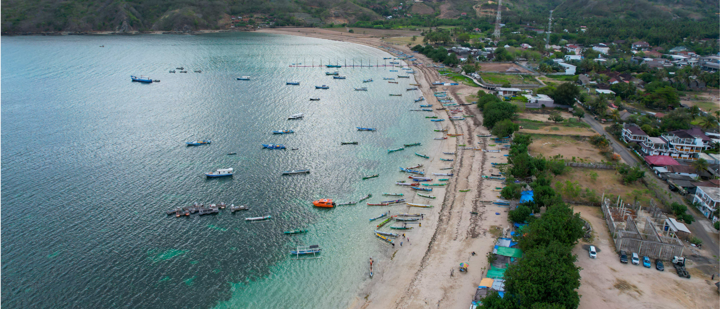 Many boats in a calm turquoise bay beside a sandy beach and coastal town, Lombok.
