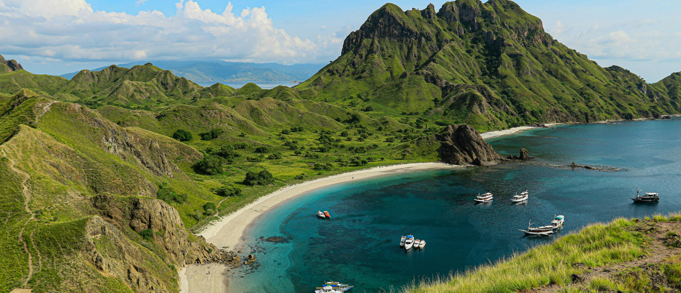 Beautiful bay with white sand beach, clear water, and boats surrounded by lush green hills.