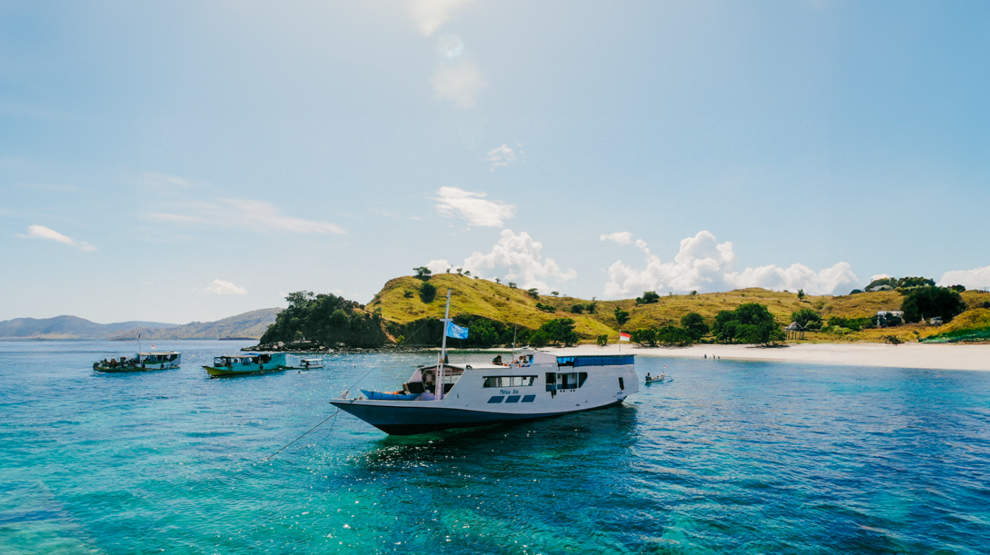 A white boat on clear turquoise water near a sunny beach and green hills in Komodo.