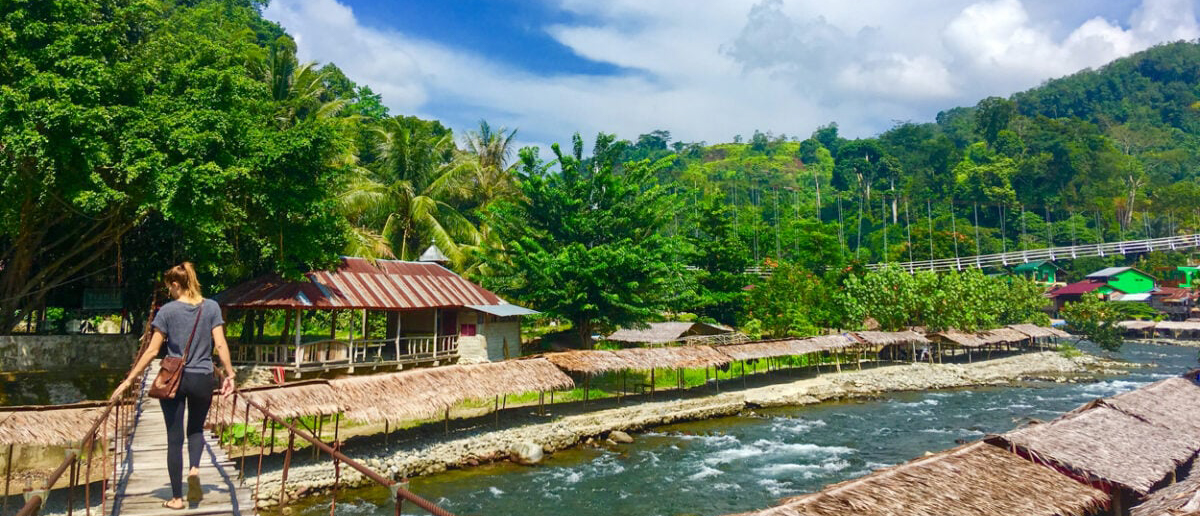 A woman walks on a wooden bridge over a river in the lush village of Bukit Lawang.