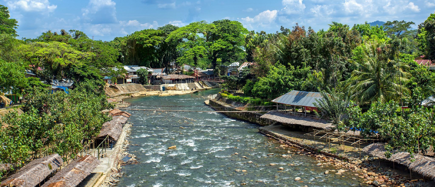 Bukit Lawang village with thatched buildings along a wide river, surrounded by dense green jungle.