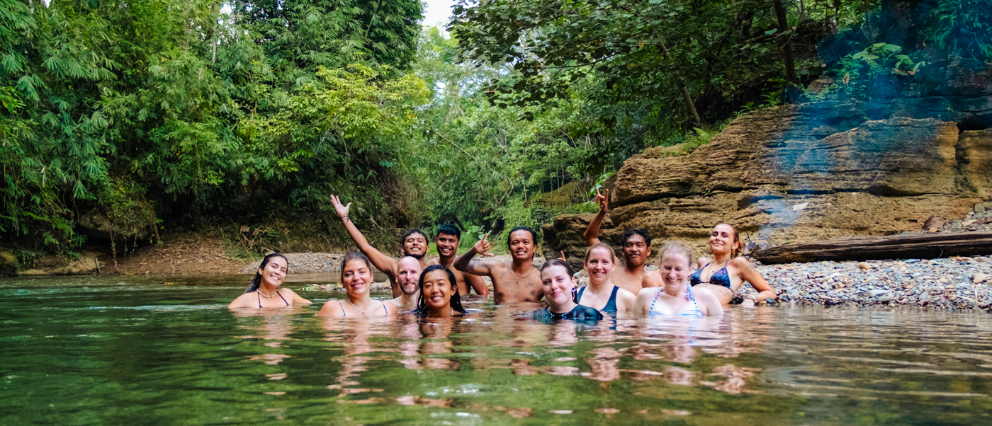 A joyful group of young travelers smiling in a clear river surrounded by lush jungle in Bukit Lawang.