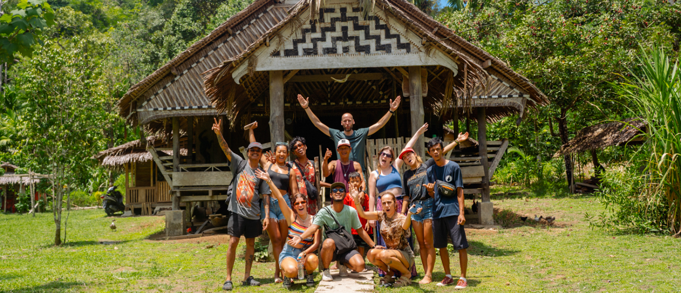A happy group of backpackers poses in front of a traditional Mentawai village hut surrounded by jungle.