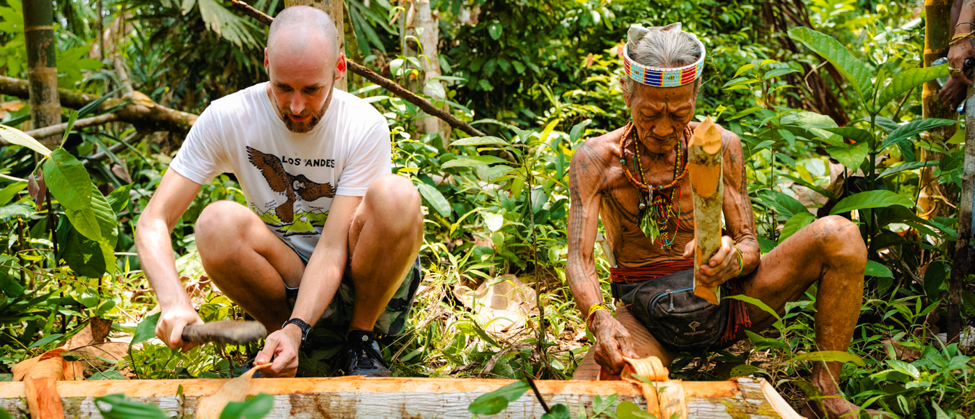 A tourist learns traditional skills from an older Mentawai tribesman in a lush Indonesian forest.