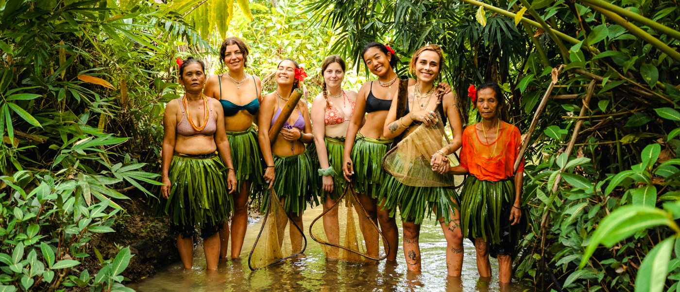 Seven women, some wearing traditional leaf skirts, smiling in a shallow Mentawai jungle stream.