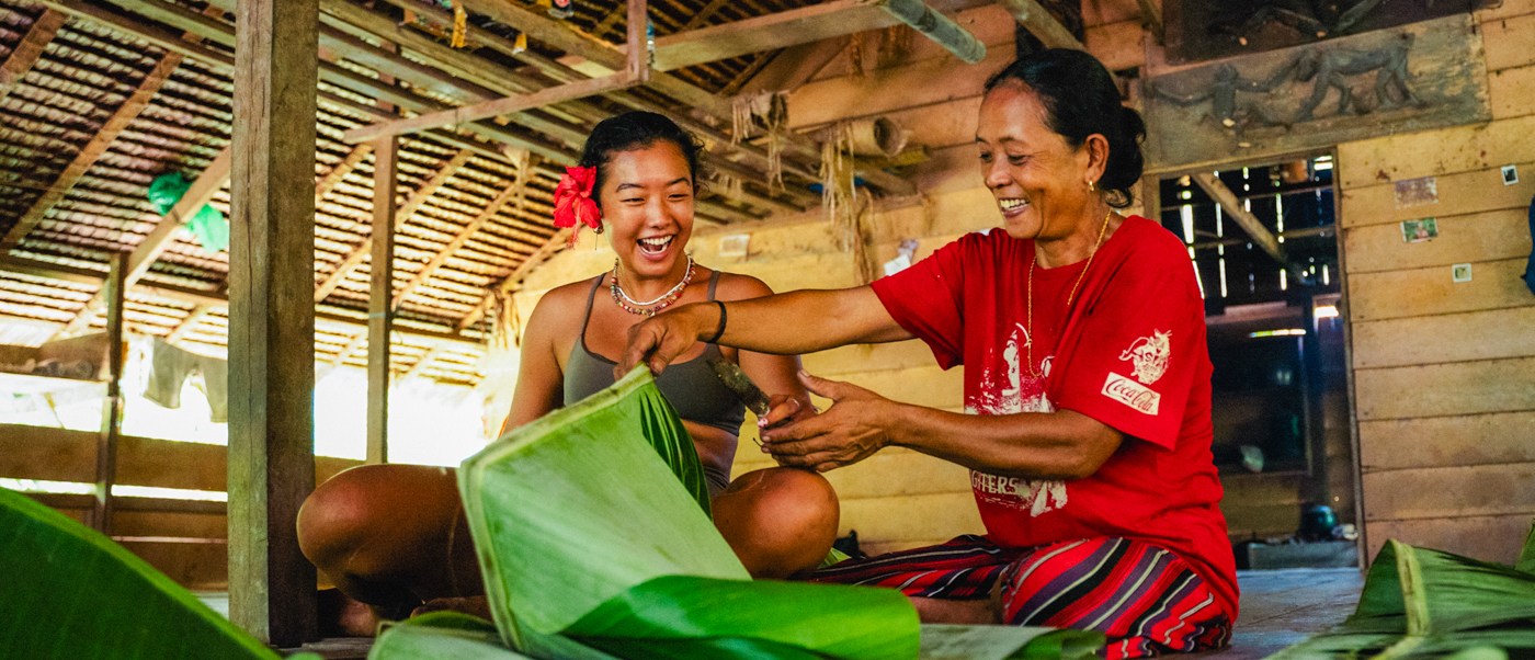 Two smiling Mentawai women inside a traditional wooden home, preparing food with green leaves.