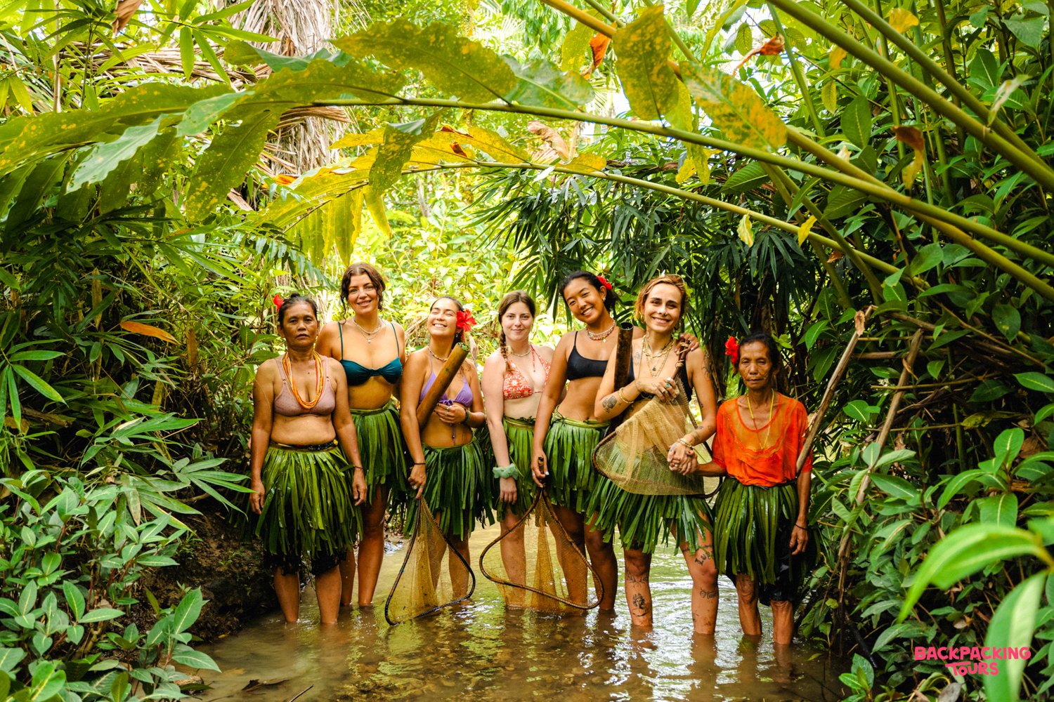 Seven women, some in traditional leaf skirts, smiling together in a shallow jungle stream in Mentawai.