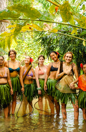 Seven women, some in traditional leaf skirts, smiling together in a shallow jungle stream in Mentawai.