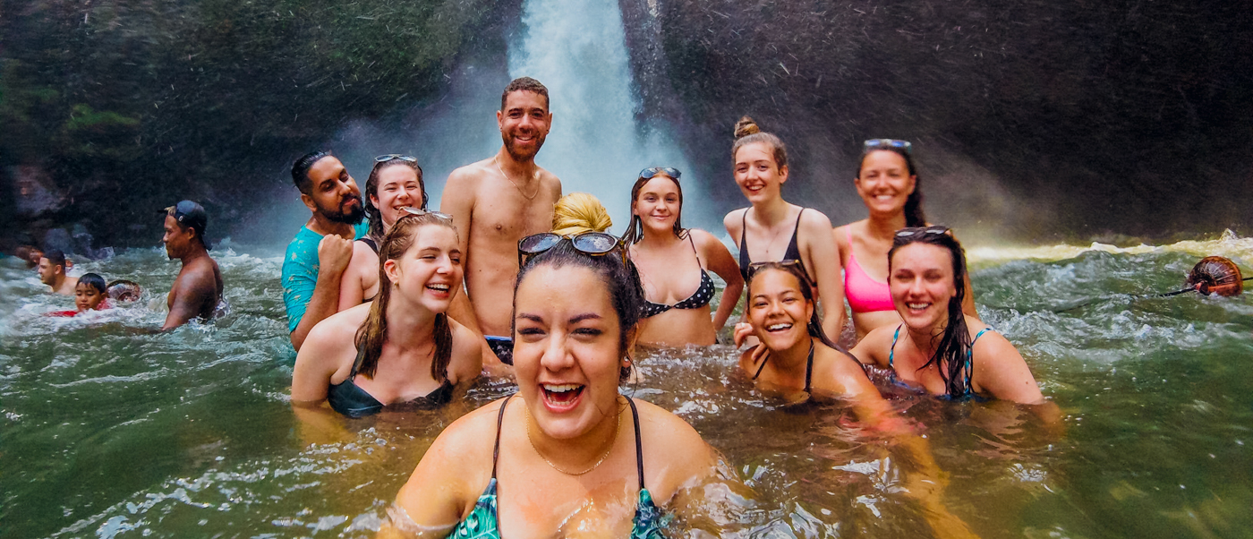 A joyful group of backpackers swimming and laughing at the base of a waterfall in Ubud, Bali.