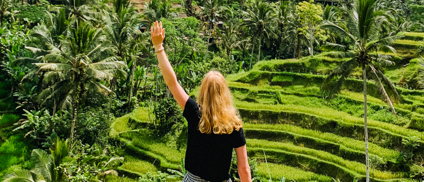 A person with blonde hair raises an arm, admiring the vibrant green rice terraces in Ubud, Bali.