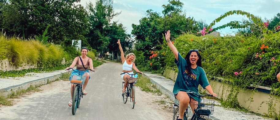 Three happy women cycle along a lush, green path on Gili Trawangan, waving excitedly.