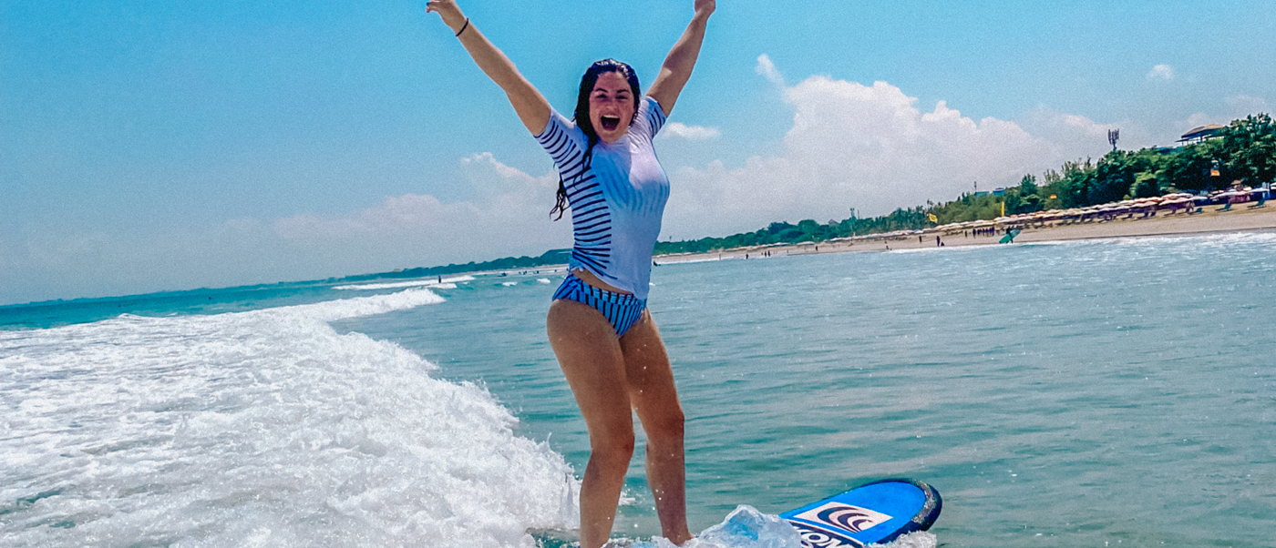 A joyful woman surfs a small wave, arms raised in excitement, on a sunny beach day.