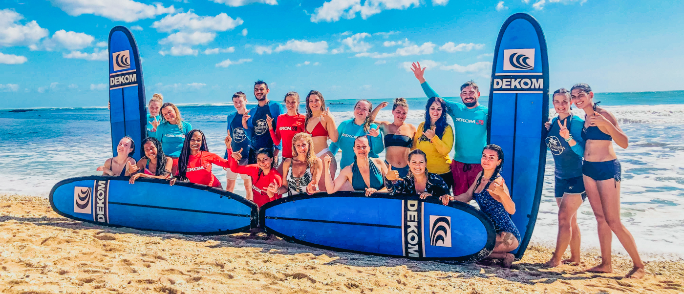 A large group of happy people pose with blue surfboards on a sunny beach in Kuta.
