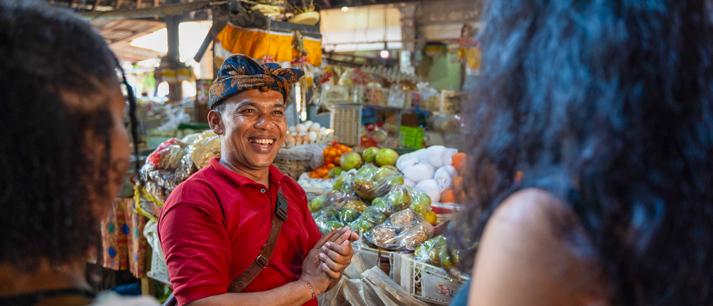 A smiling local Balinese man in traditional headwear talks to tourists at a bustling Ubud market.