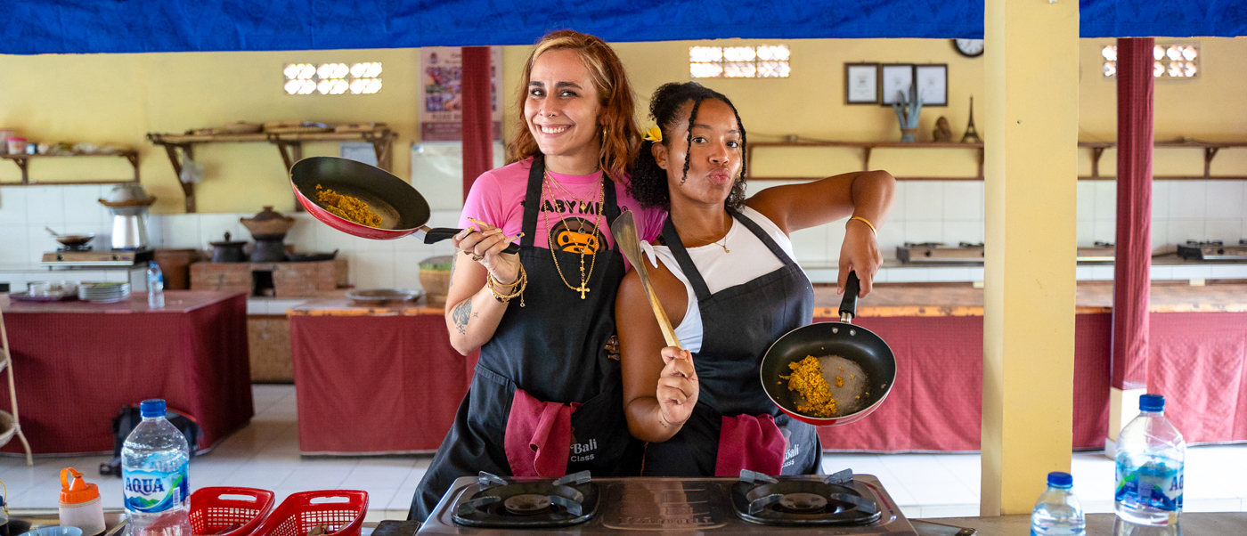 Two joyful women cooking and tossing food in pans during a class in Ubud.