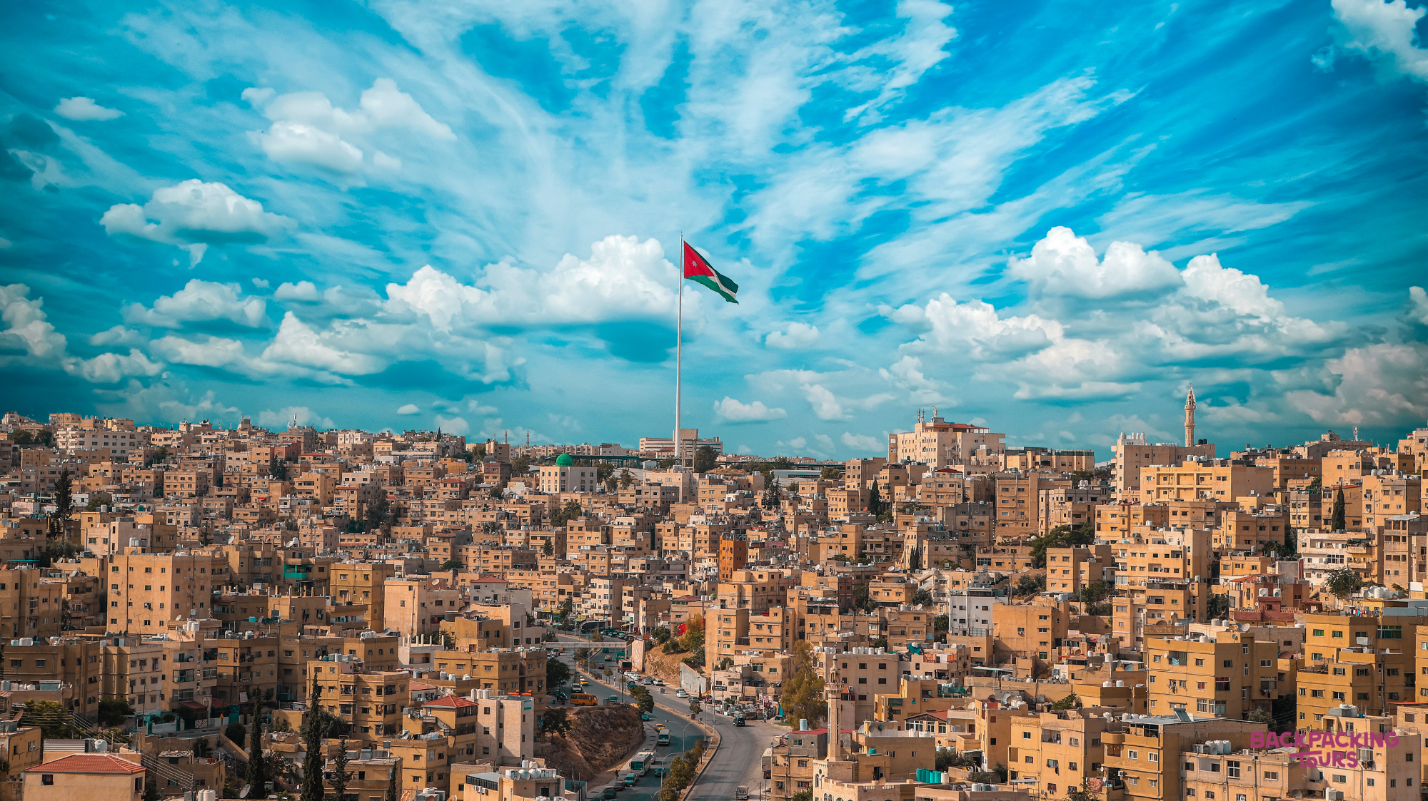 The Jordanian flag proudly flies over Amman's sprawling cityscape of light-colored buildings under a vibrant sky.