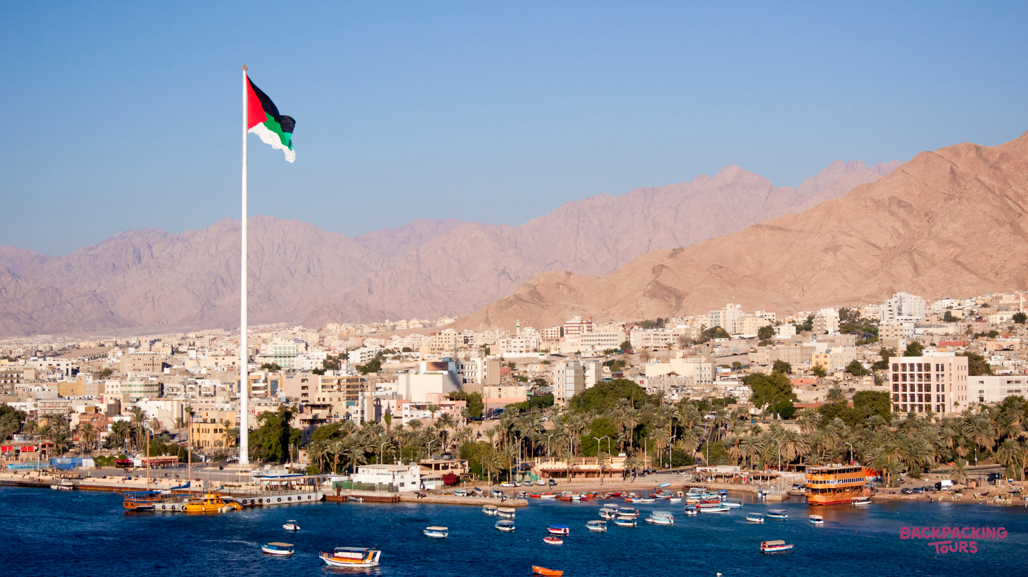 Aqaba's bustling waterfront with boats on the Red Sea, the Jordanian flag, and arid mountains.
