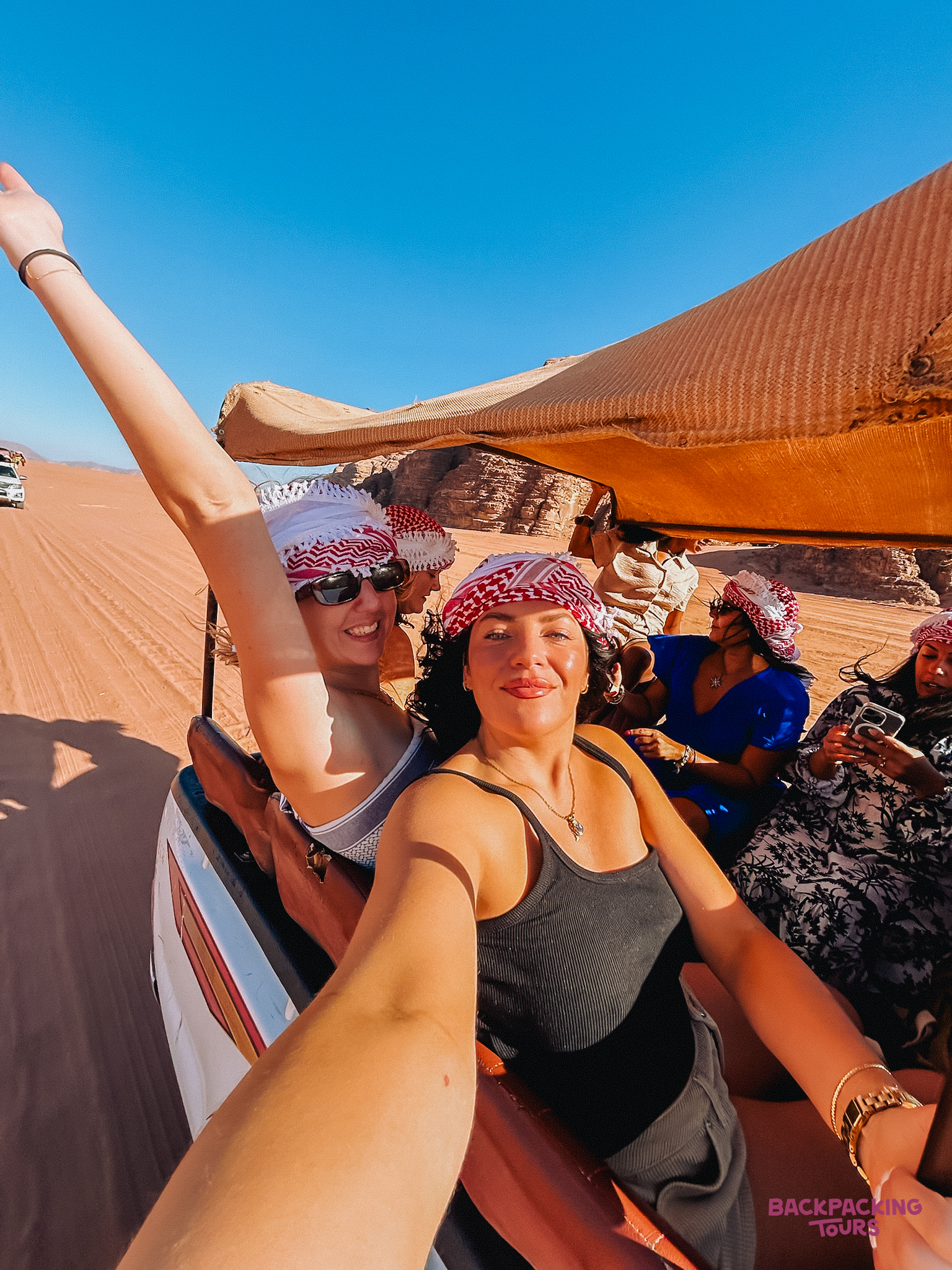 Backpackers smiling during an adventurous jeep ride across the vibrant red sands of Wadi Rum.