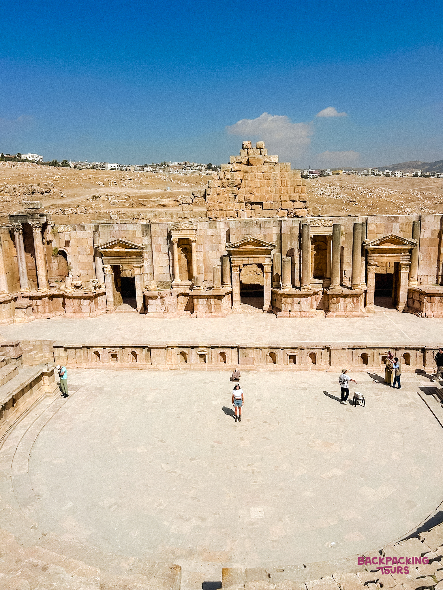 Tourists explore the grand ancient Roman amphitheatre at Jerash under a clear blue sky.
