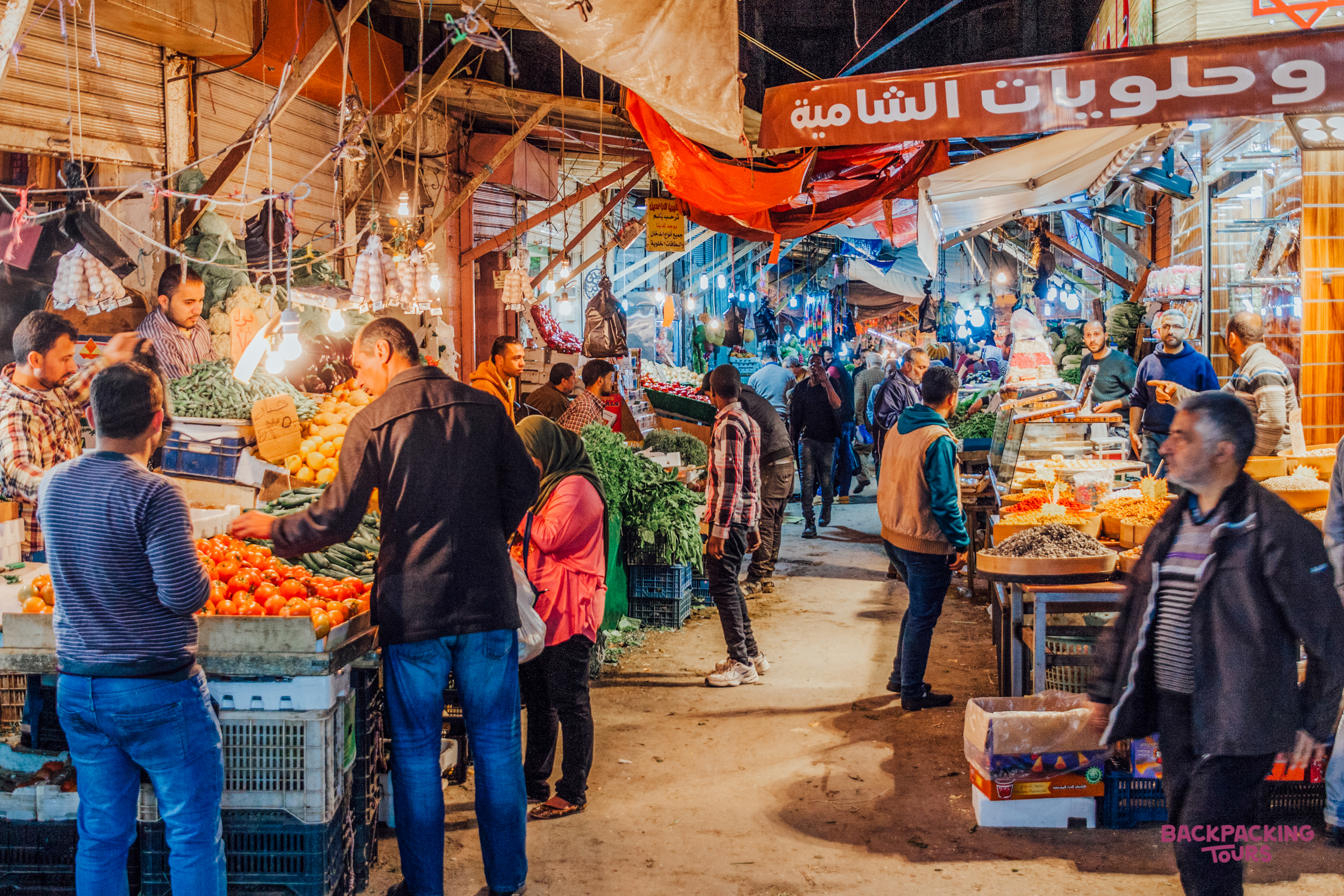 Shoppers browse vibrant produce stalls at a bustling market in Amman, Jordan, under warm lights.