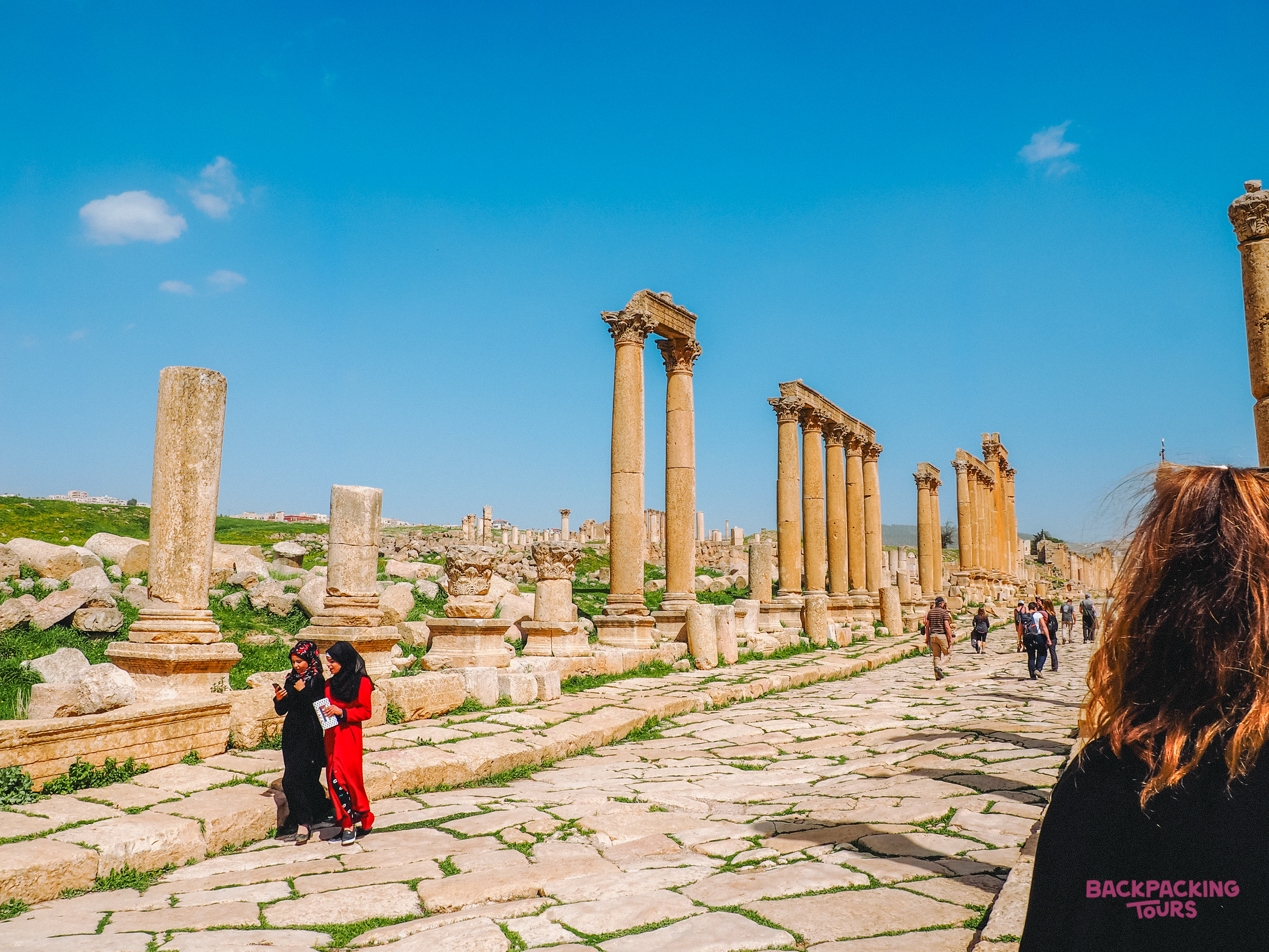 Visitors wander through the impressive ancient Roman ruins of Jerash, Jordan, under a clear blue sky.