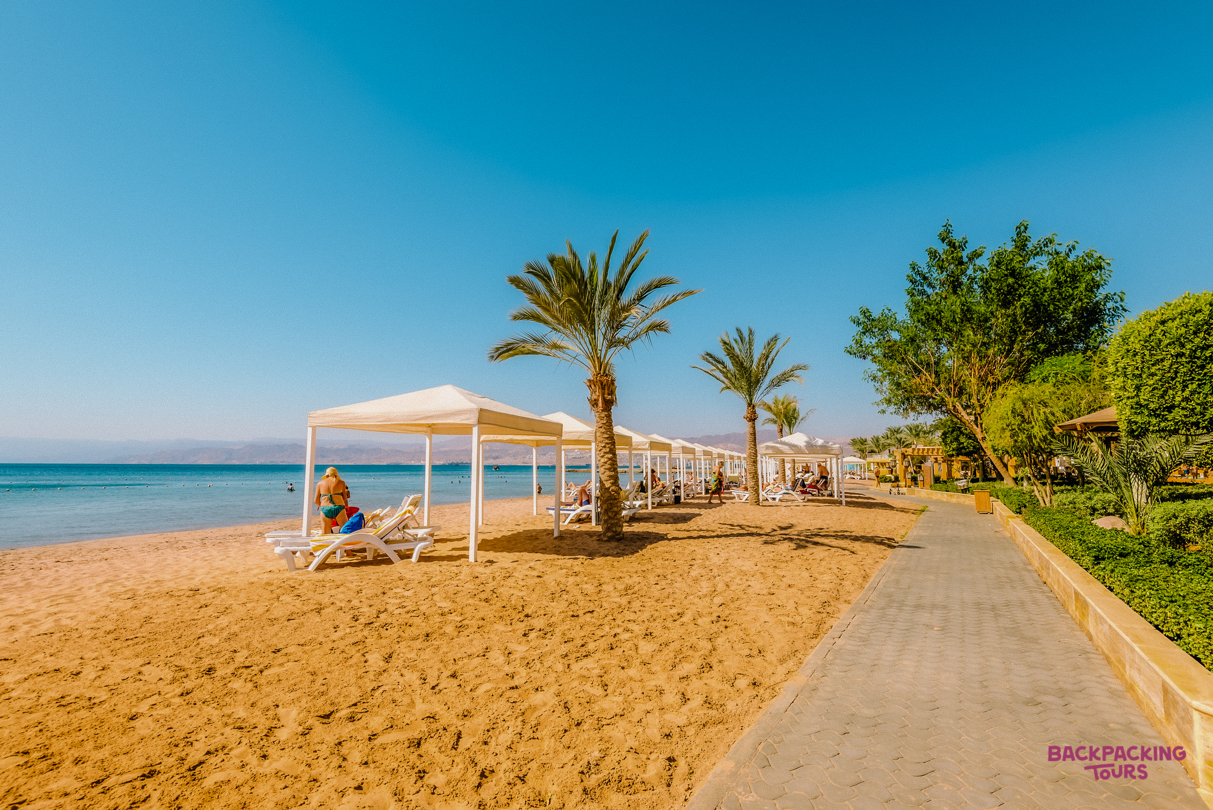Tourists relax under white canopies on a sunny Dead Sea beach with calm blue water.