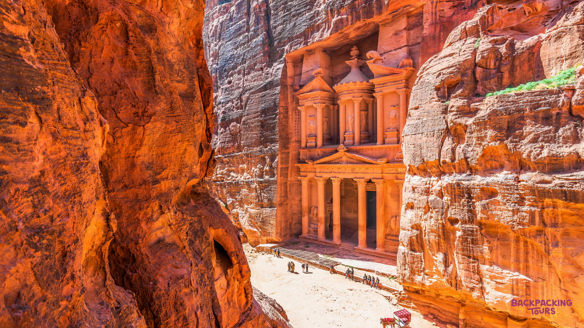 The magnificent Treasury of Petra emerges dramatically from the narrow canyon, with small figures exploring.