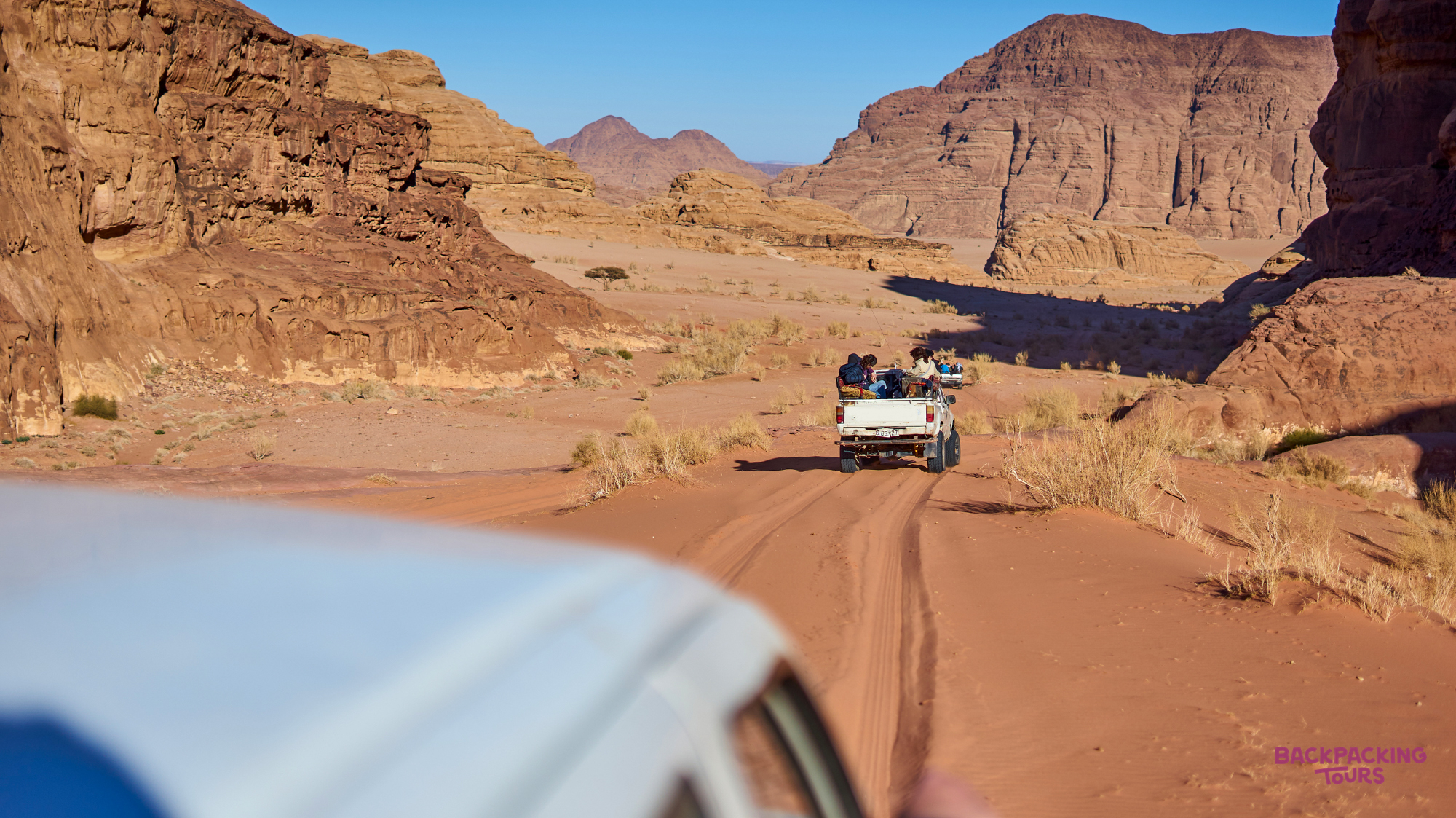 A white jeep carrying passengers drives along a sandy track through the majestic Wadi Rum desert.