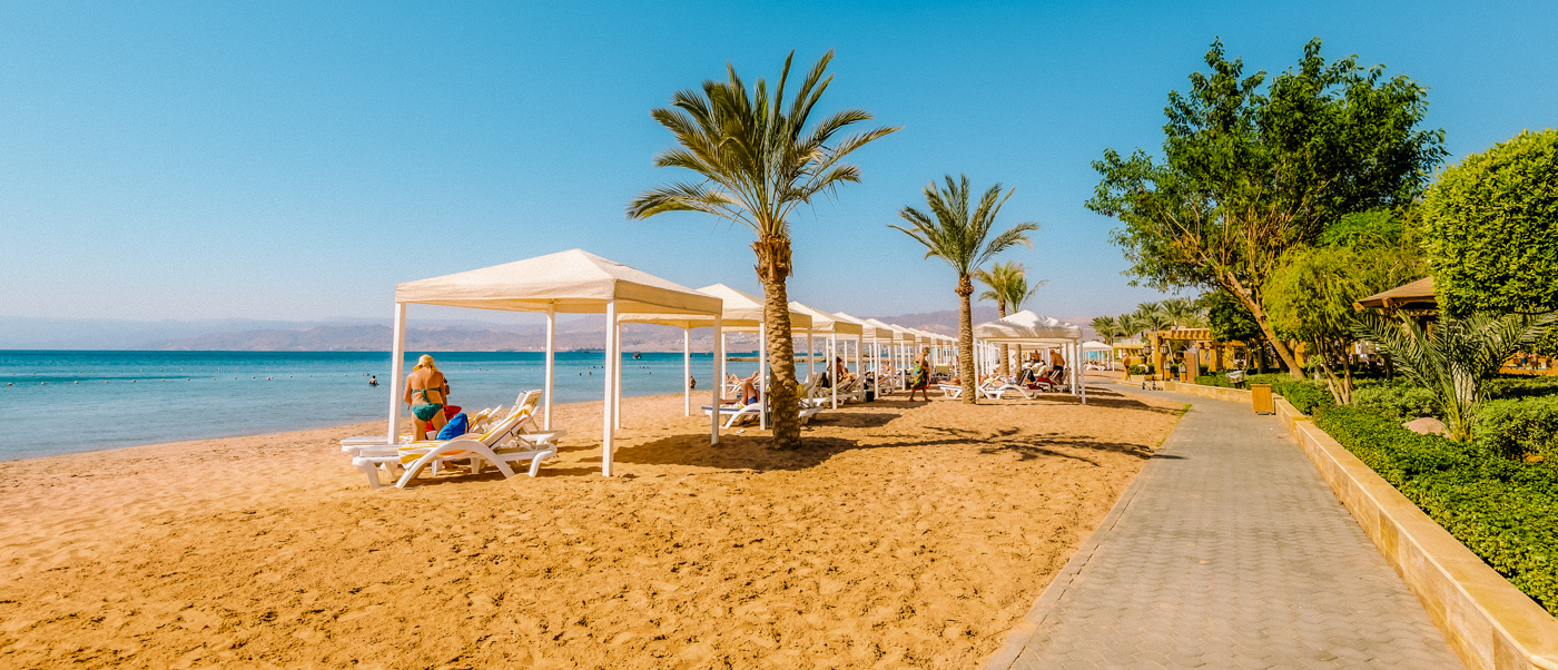 People relax under white canopies on a sunny sandy beach with palm trees by the Dead Sea.