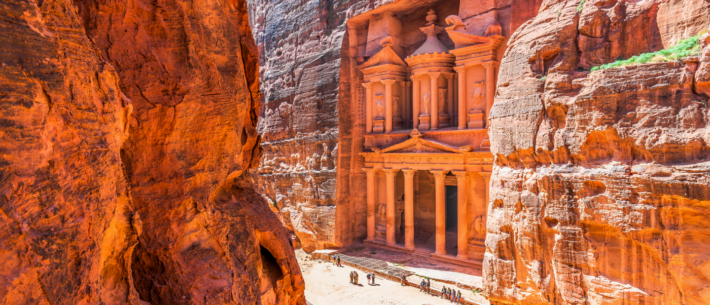 Tourists stand dwarfed by the magnificent rock-cut Treasury of Petra, emerging from a narrow canyon.