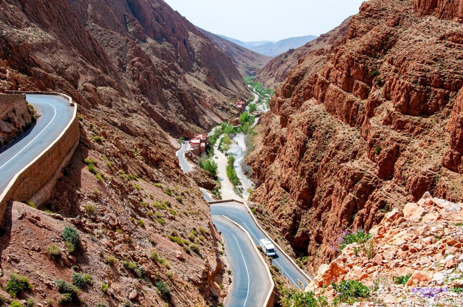 A winding road follows a green river valley between steep red rock walls near Todra Gorge.