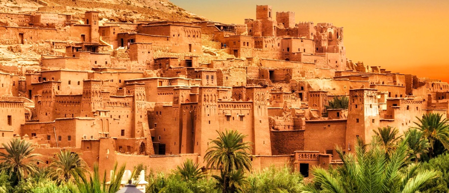 The earthen towers and walls of Ait Benhaddou glow orange above palm trees at sunset.