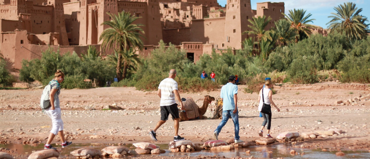 Travellers cross a shallow river on stepping stones below the mud-brick kasbah of Ait Benhaddou.