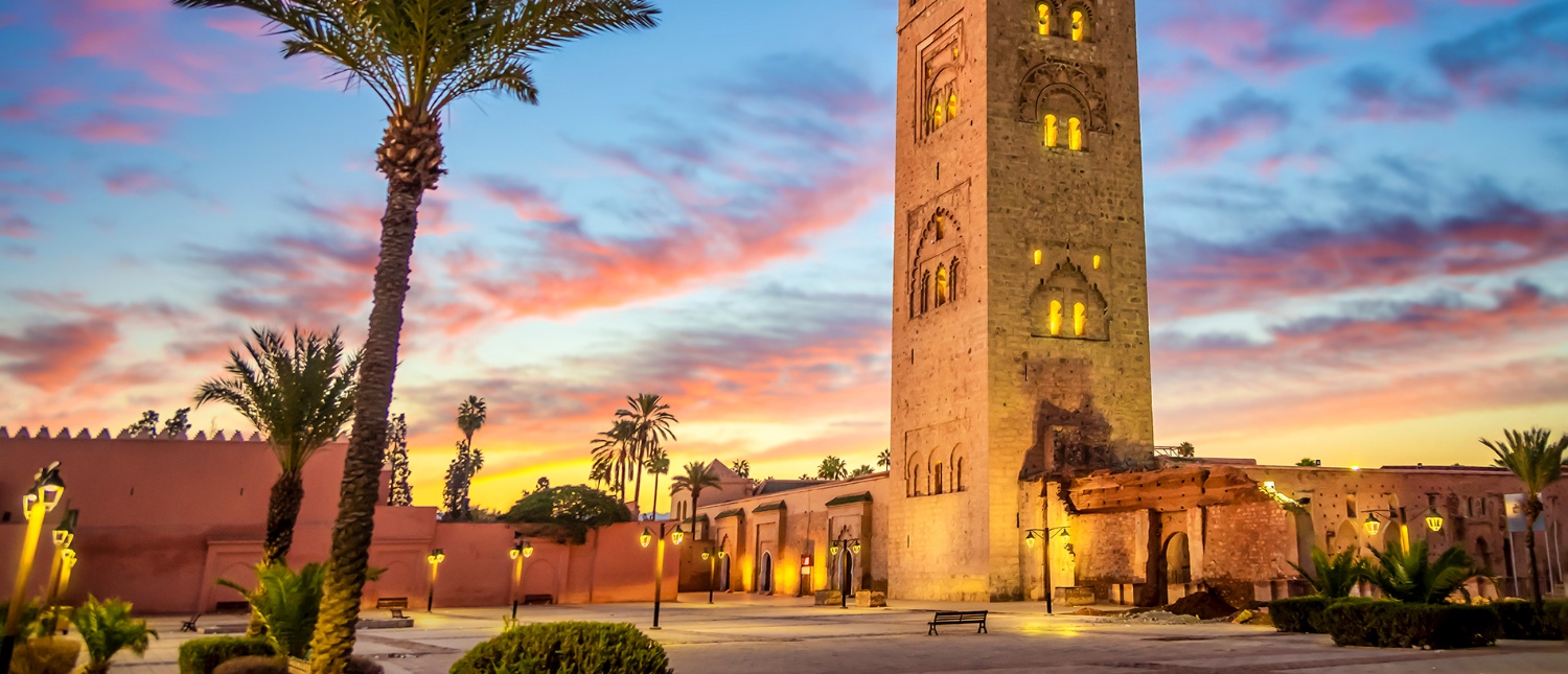 The Koutoubia Mosque minaret and palm trees against a pink and orange sunset in Marrakech.