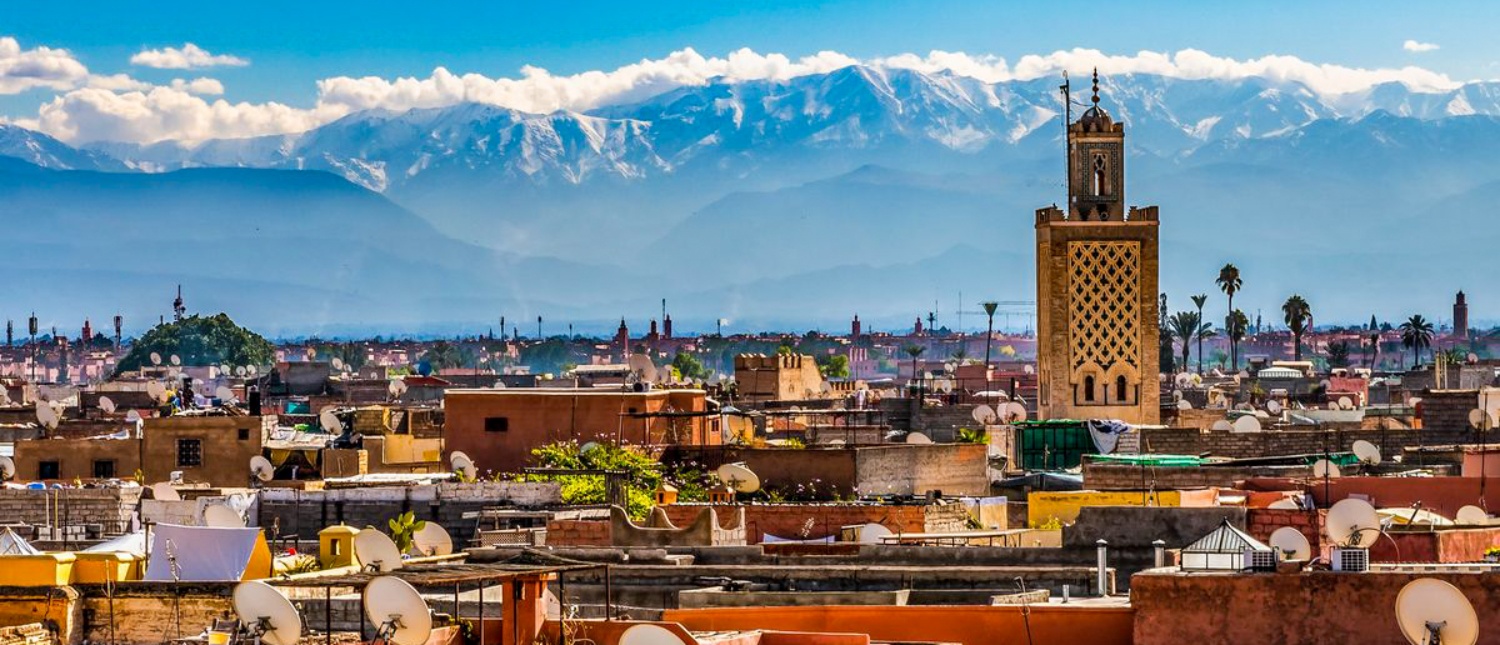 Marrakech rooftops and a tall minaret, with the snow-capped High Atlas rising behind the city.