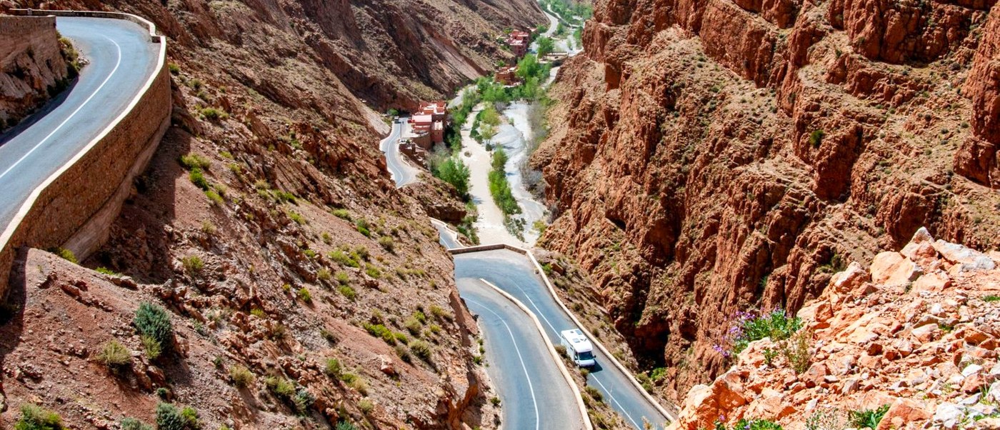 A winding road follows a green river valley between steep red rock walls near Todra Gorge.