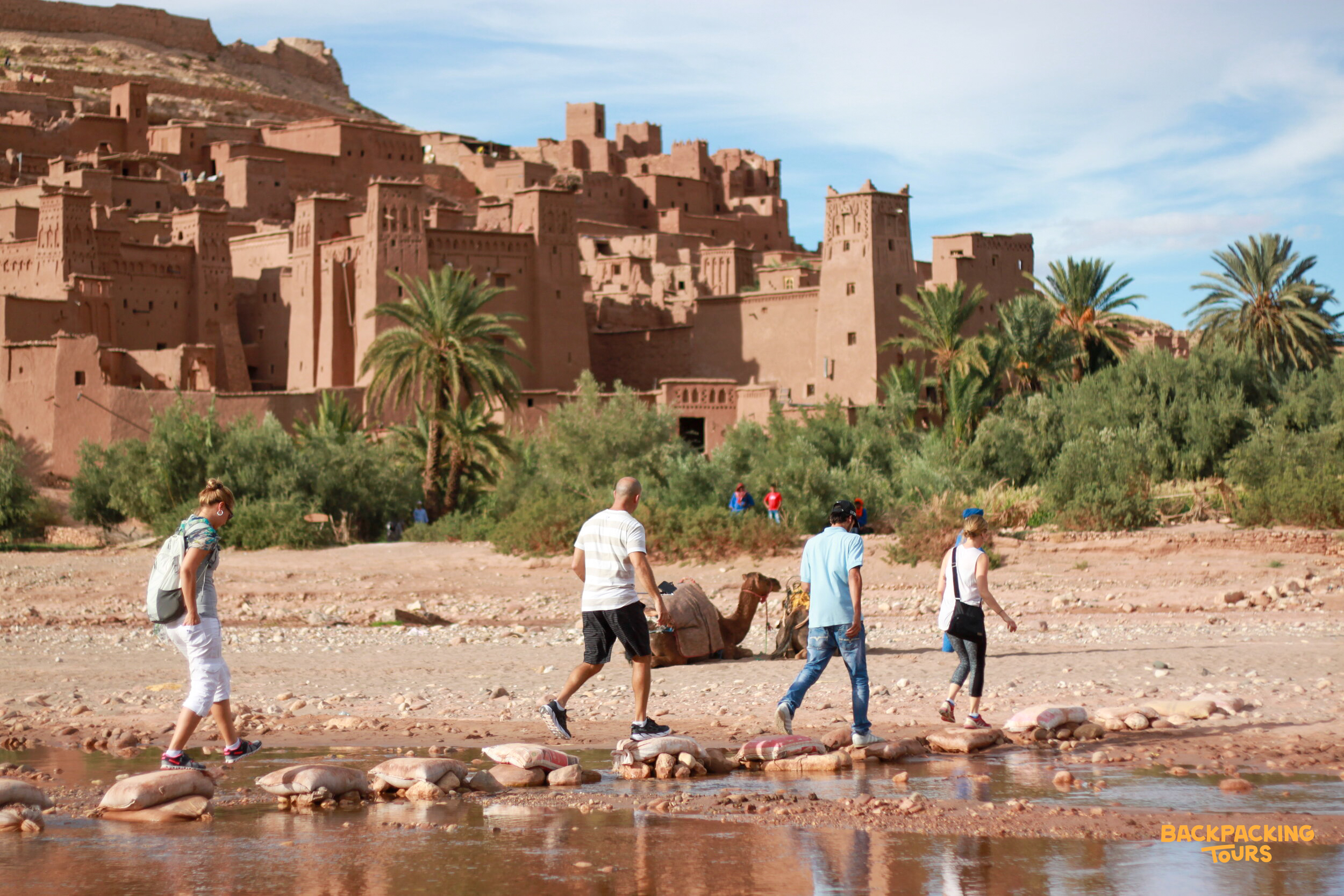 Four travelers cross a river on stepping stones with the historic Ait Ben Haddou Kasbah in Morocco.