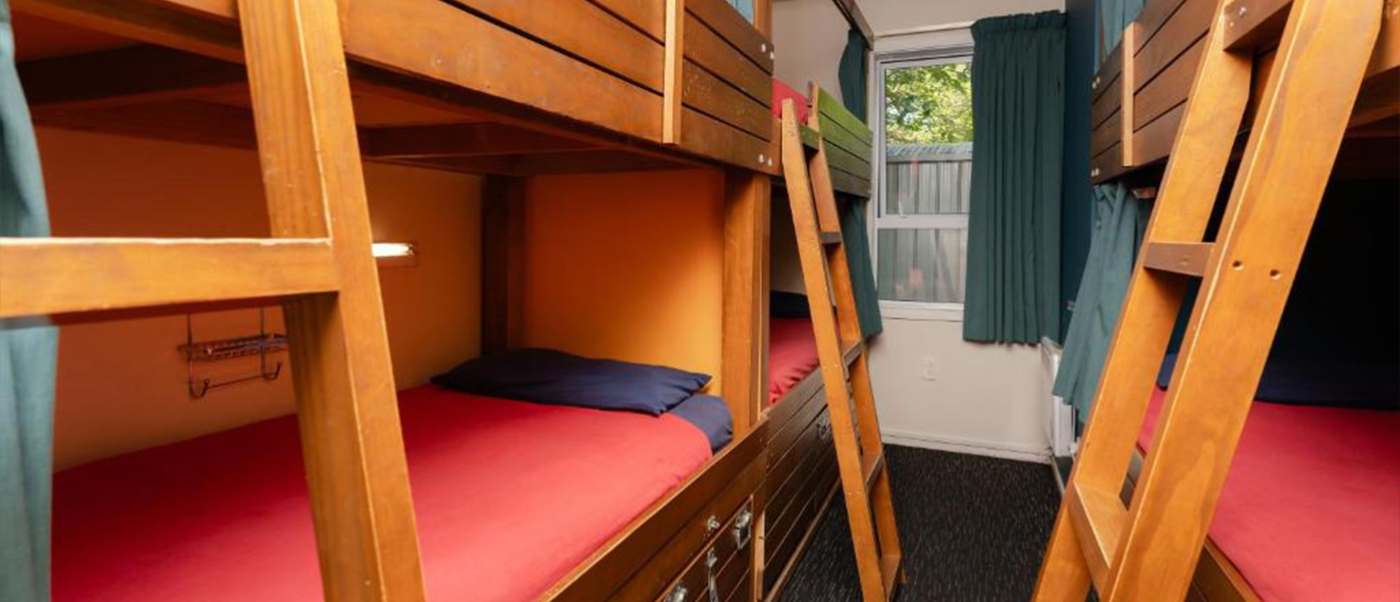 A clean hostel dorm room in Queenstown, New Zealand, with multiple wooden bunk beds and red bedding.